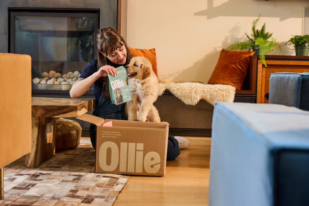 A woman sitting on the floor next to an Ollie box with a Golden Retriever in a bright living room.