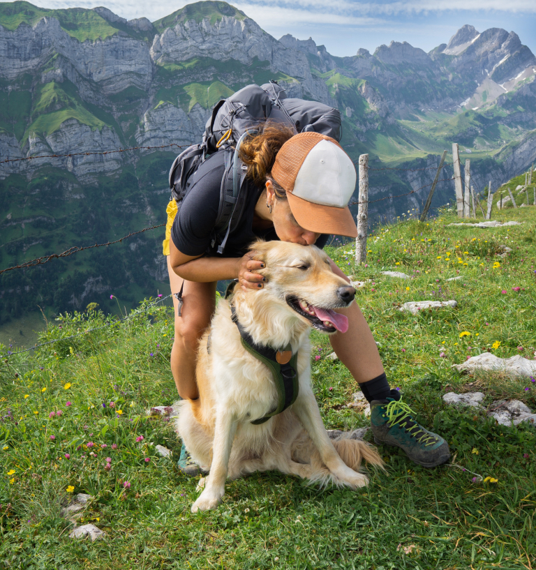 A woman in hiking gear kissing her golden retriever's head while they stand on a ridge of mountains with rocky mountains in the background.